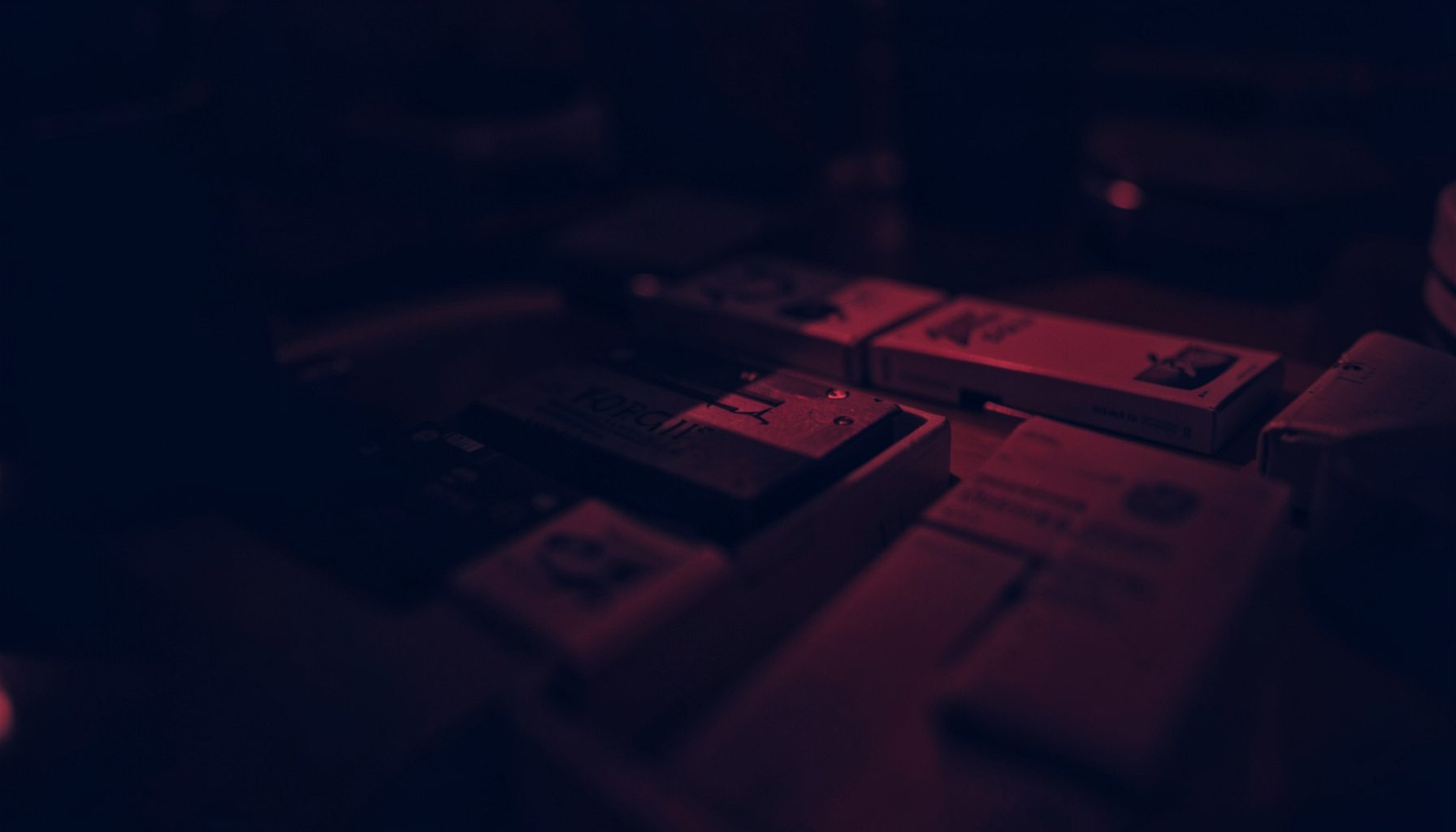 Dark atmospheric duotone photograph in deep navy and muted crimson, showing a close-up of cigarette packs arranged on a reflective surface with soft shadows and moody lighting.